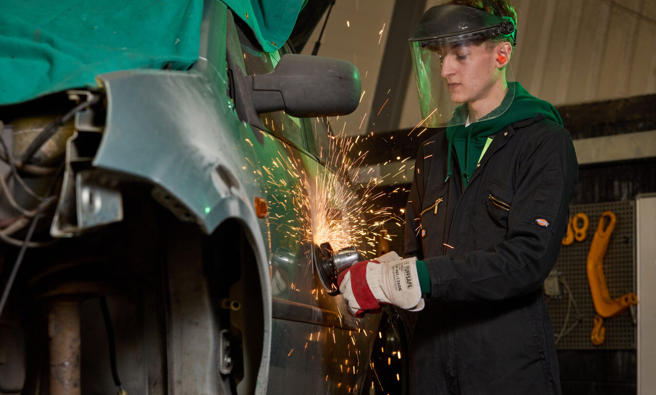 Photo of a young man wearing protective clothing and using a grinder on the side of a motor vehicle