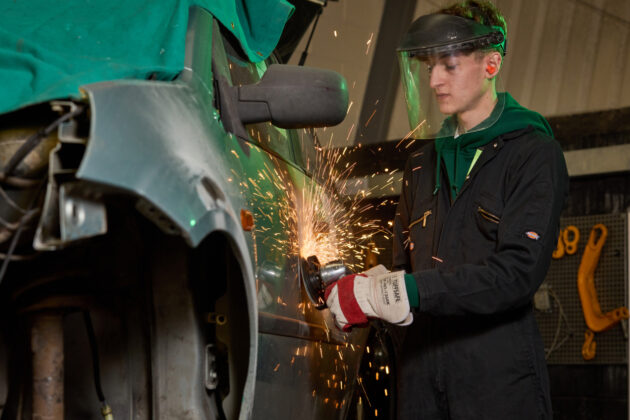 Photo of a young man wearing protective clothing and using a grinder on the side of a motor vehicle