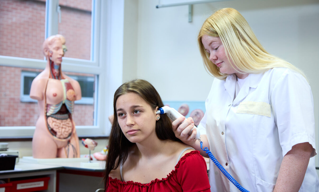 Photo of a student checking the temperature of another student with an ear thermometer