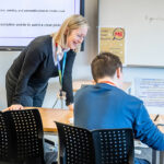 Looking at the back of a student who is sat at a desk, a tutor stood supporting them