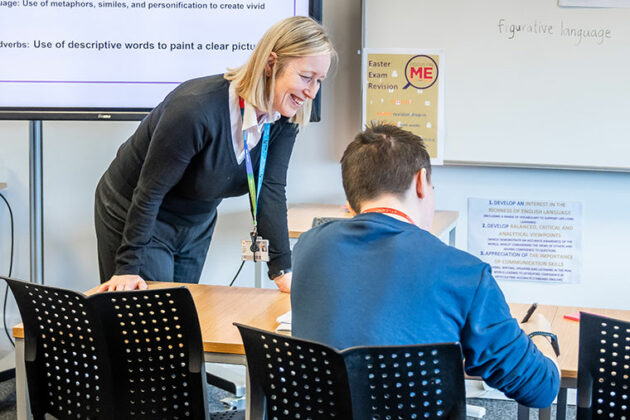 Looking at the back of a student who is sat at a desk, a tutor stood supporting them