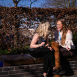 Two smiling students chatting sat on a wall with bushes behind them
