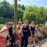Six people stood in a line against a fence with trees and a blue sky in the background