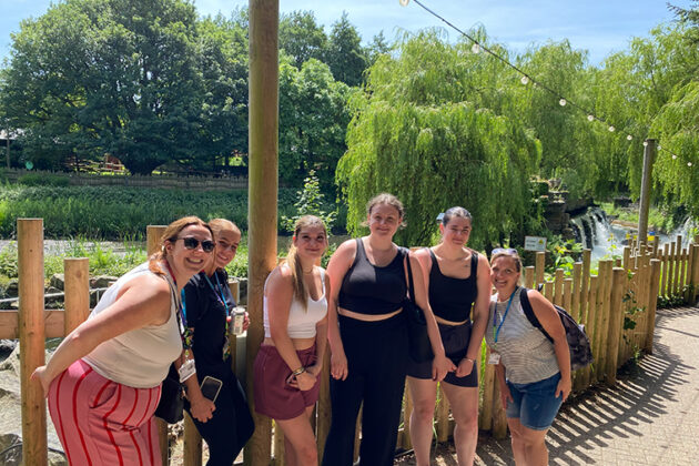 Six people stood in a line against a fence with trees and a blue sky in the background