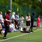 Three people stood at the start line about to start an egg and spoon race, a crowd of people behind them watching