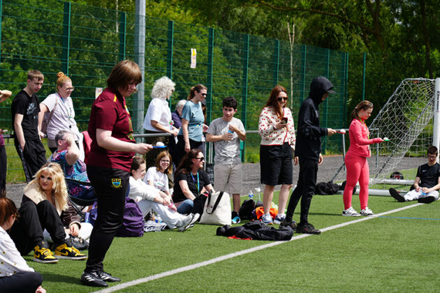 Three people stood at the start line about to start an egg and spoon race, a crowd of people behind them watching