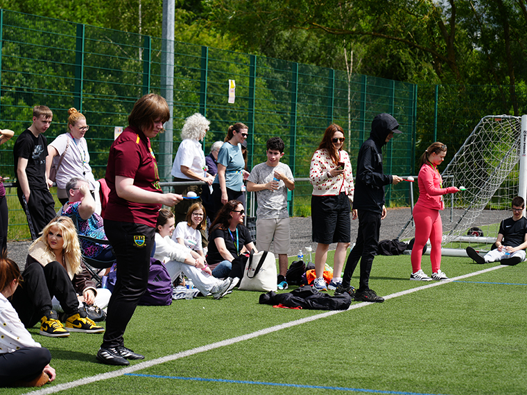 Three people stood at the start line about to start an egg and spoon race, a crowd of people behind them watching