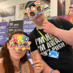Two people holding up rainbow coloured cardboard glasses up to their faces
