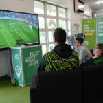Three students sat on a black sofa looking at a large screen with a football game on