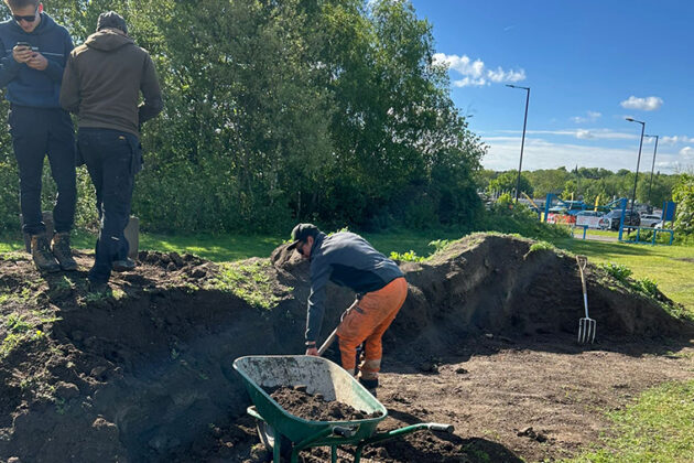 Someone in orange trousers and a blue top digging a large hole in the grass, a wheel barrow at the side of them