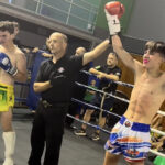 Two boxers stood in a boxing ring with a referee stood between them holding one of the boxer's arms up