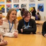 Four smiling students sat together chatting and leaning on a table, artwork up on the wall in the background