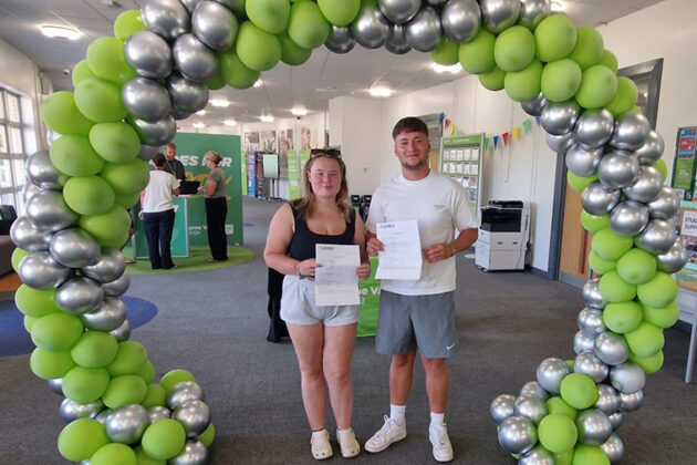 A male and a female stood under a green and silver balloon arch, smiling and each holding a piece of paper