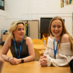 Two smiling students sat at a desk chatting to friends