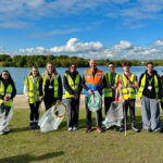 A group of people stood next to a lake, each wearing high vis jackets and holding bags and litter pickers