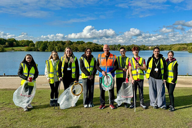 A group of people stood next to a lake, each wearing high vis jackets and holding bags and litter pickers