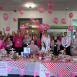 A large crowd of people mostly wearing pink, stood behind a long table with a red and white checked table cloth on, lots of pink balloons in the air