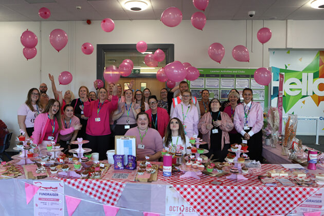 A large crowd of people mostly wearing pink, stood behind a long table with a red and white checked table cloth on, lots of pink balloons in the air